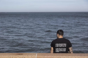 de espaldas un hombre joven mira el rio de la plata sentado en la rambla. tiene una camiseta con el siguiente texto: la guerra la mató sin darle un balazo, a la guerra le dan miedo los abrazos, la guerra con camuflaje se viste así nadie ve cuando se pone triste. La guerra pierde todas las luchas cuando los enemigos se escuchan, la guerra es más fácil que fuerte, no aguanta la vida por eso se esconde  en la muerte