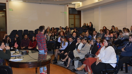 Fotografía durante la actividad. Se aprecia el auditorio con egresados/as y sus familiares y el Docente Bruno Cancio de espaldas tomando la palabra.