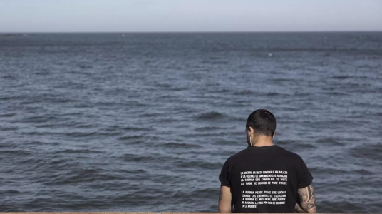 de espaldas un hombre joven mira el rio de la plata sentado en la rambla. tiene una camiseta con el siguiente texto: la guerra la mató sin darle un balazo, a la guerra le dan miedo los abrazos, la guerra con camuflaje se viste así nadie ve cuando se pone triste. La guerra pierde todas las luchas cuando los enemigos se escuchan, la guerra es más fácil que fuerte, no aguanta la vida por eso se esconde  en la muerte